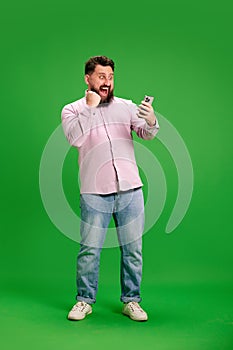 Young bearded man looking at smartphone with victorious expression and clenched fist of success against vibrant green