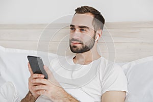 Young bearded man lying in bed with white sheet pillow blanket in bedroom at home. Male spending time in room using