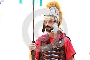 A young bearded man in a red uniform of a Roman legionary, in a metal helmet, tunic and with a spear stands in