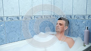 Young bearded man is relaxing lying in bath with foam at home after busy day.