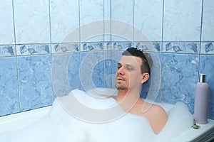 Young bearded man is relaxing lying in bath with foam at home after busy day.