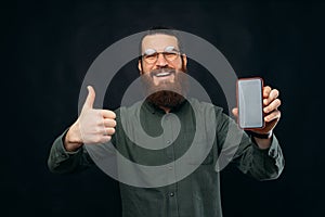 Young bearded man is showing to the camera blank screen phone a a thumb up.