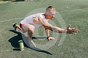 Young bearded man sitting on basketball court outdoors with ball holding bottle drinking electrolytes drink.