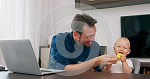 Young bearded man sitting at desk at home with laptop Cute baby sits at high baby chair, father holds teether which baby