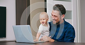 Young bearded man sitting at desk at home with laptop, holding cute baby at knees, talking via communicator to baby