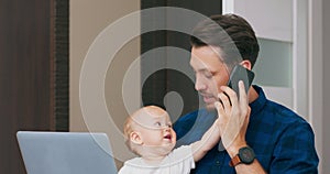 Young bearded man sitting at desk at home with laptop, holding cute baby at knees, talking via smartphone Baby looks at