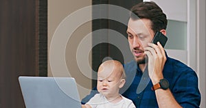Young bearded man sitting at desk at home with laptop, holding cute baby at knees, talking via smartphone Baby touches