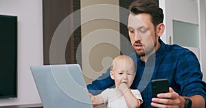 Young bearded man sitting at desk at home with laptop, holding cute baby at knees, writing on keyboard, reading message