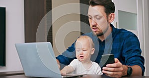 Young bearded man sitting at desk at home with laptop, holding cute baby at knees, writing on keyboard, reading message