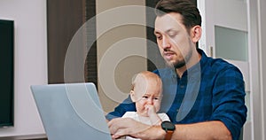 Young bearded man sitting at desk at home with laptop, holding cute baby at knees, writing on keyboard, talking to baby