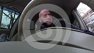 Young bearded man sitting in his car in the driver's seat. He is starting driving and looking at the camera.