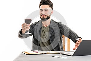 Young bearded man sitting by the table holding a glass of wine in his hand and cover the web cam