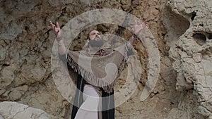 A young bearded man with a staff in his hands in the clothes of a shaman prays on a rock among the sandy dunes of the