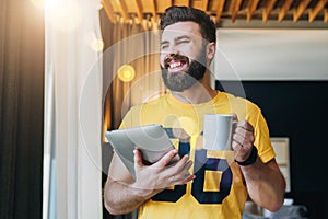 Young bearded man stands in room and holding tablet computer while drinking coffee. Guy freelancer working at home.