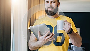 Young bearded man stands in room and holding tablet computer while drinking coffee. Guy freelancer working at home.