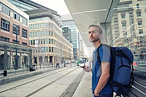Young bearded man tourist with backpack and smartphone stand on public transport stop and waiting tram in modern city center.