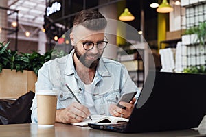 Young bearded man trendy glasses sits cafe in front of laptop computer, uses smartphone, takes notes in notebook