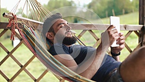 A young bearded man uses a tablet, lying in a hammock in a country house