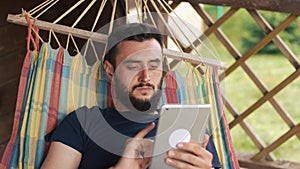 A young bearded man uses a tablet, lying in a hammock in a country house