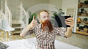 Young bearded man using tablet computer having video chat sitting in bed at home