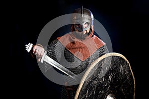 Young bearded man in a Viking-era helmet covering his face holding a sword and shield