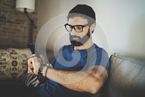 Young bearded man wear casual clothes and eye glasses checking time on smartphone screen at modern loft home.Horizontal