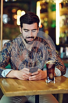 Young bearded man wearing casual clothes looking at his smartphone in a modern pub