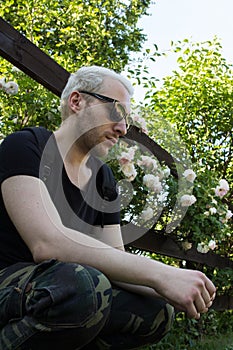 A young bearded man wearing sunglasses by the fence of blooming garden. A guy sitting by the fence and looking down.