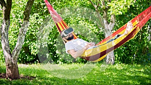 Young bearded man wearing virtual reality goggles relaxing in a garden hammock. Lifestyle VR fun and relax