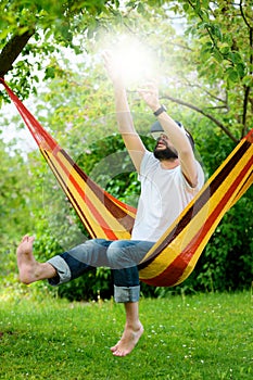 Young bearded man wearing virtual reality goggles relaxing in a garden hammock. Lifestyle VR fun