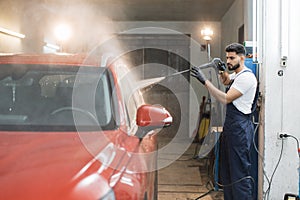 Young bearded man worker washing red crossover car under high pressure water in car wash service.