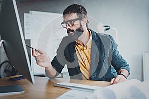 Young bearded man working at sunny office on desktop computer while sitting at wooden table.Businessman analyze stock