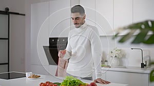 Young bearded Middle Eastern man smelling healthful fresh tomato smiling standing in kitchen at home. Positive confident