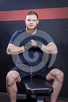 Bearded guy, a mixed martial arts fighter, sits on a couch, brought his hands together in front of his chest. Looks at the camera.