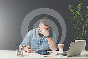 Young bearded sad businessman is sitting at table, covering his face with his hand and holding smartphone in his hand.