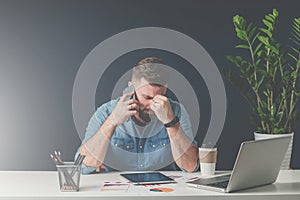 Young bearded sad businessman is sitting at table, covering his face with his hand and talking on cell phone.