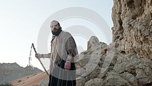 A young bearded shaman with a staff in his hand walks along the rocks in the middle of the desert