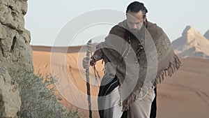 A young bearded shaman with a staff in his hand walks along the rocks in the middle of the desert