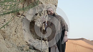 A young bearded shaman with a staff in his hand walks along the rocks in the middle of the desert