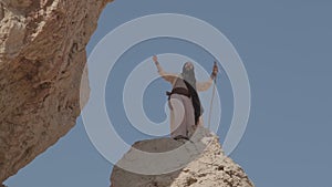 A young bearded shaman with a staff in his hands, in white clothes, prays standing on a rock against the background of a