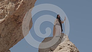 A young bearded shaman with a staff in his hands, in white clothes, prays standing on a rock against the background of a