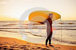 Young bearded surfer in wetsuit with yellow surfing longboard on a ocean coast at sunset. Water sport adventure camp and