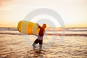 Young bearded surfer in wetsuit with yellow surfing longboard walk from water at sunset ocean. Water sport adventure camp and