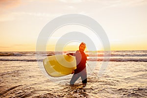 Young bearded surfer in wetsuit with yellow surfing longboard walk from water at sunset ocean. Water sport adventure camp and