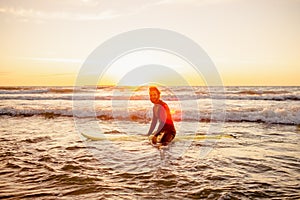 Young bearded surfer in wetsuit with yellow surfing longboard in a water at sunset ocean. Water sport adventure camp and extreme