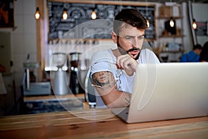 Young bearded tattooed guy in white t-shirt sitting behind the table in bar