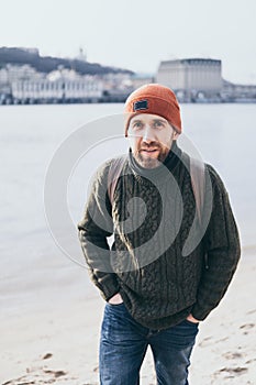 Young bearded traveller with rucksack standing at the river front with cityscape on background