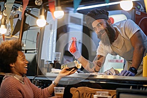 Young beardy caucasian employee in fast food adding a ketchup in a sandwich to a female afro-american customer, joking