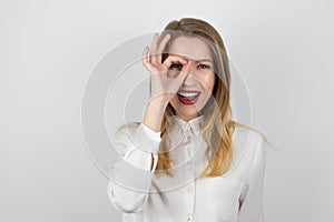 Young beatuiful blond woman holding her hand like magnifying glass near her eye looks surprised, splash of emotions