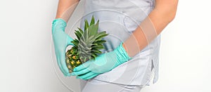 Young beautician wearing blue gloves in uniform with pineapple covers an intimate area on a white background, bikini